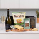 The Sparkling Picnic Treat Box. A display of gourmet food items arranged on a wooden platter. The arrangement includes a bottle of sparkling wine, a bag of Keogh's Irish cheese & onion crisps, a block of cheese, a box of chocolate biscuit bars, and a package of Irish oat cookies. A glass filled with sparkling wine is positioned nearby. The Sparkling Picnic Treat Box. A display of gourmet food items arranged on a wooden platter. The arrangement includes a bottle of sparkling wine, a bag of Keogh's Irish cheese & onion crisps, a block of cheese, a box of chocolate biscuit bars, and a package of Irish oat cookies. A glass filled with sparkling wine is positioned nearby.